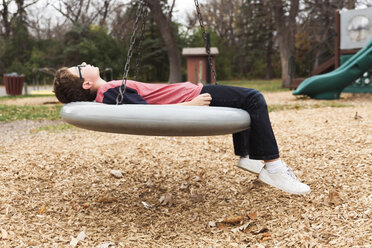 Side view of boy lying on tire swing at playground - CAVF49806