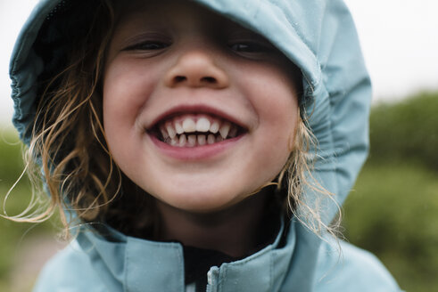 Close-up portrait of cute happy girl in blue raincoat standing against plants during rainy season - CAVF49815