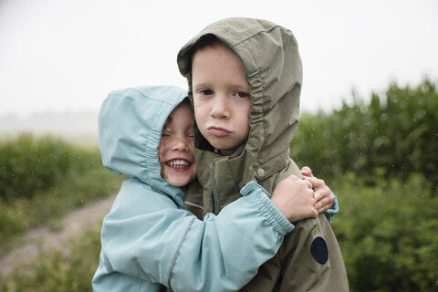 Portrait of sad brother being embraced by happy sister while standing against plants during rainy season stock photo