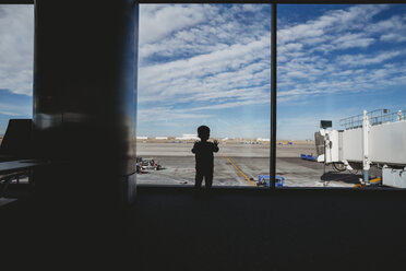Rear view of silhouette baby boy looking through window at airport departure area - CAVF49866