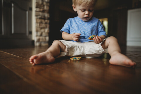 Niedlicher kleiner Junge isst Keks, während er zu Hause auf dem Boden sitzt, lizenzfreies Stockfoto