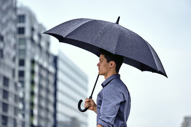 Seitenansicht eines jungen Geschäftsmannes mit Regenschirm, der vor Gebäuden und dem Himmel in einer Stadt steht, während es regnet - CAVF49896