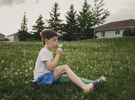 Side view of boy with broken leg blowing dandelion while sitting on grassy field at park - CAVF49909