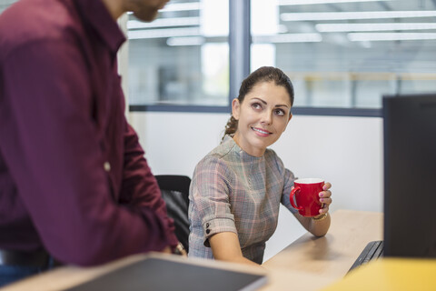 Weibliche Angestellte sitzt am Schreibtisch und spricht mit einem Kollegen, lizenzfreies Stockfoto