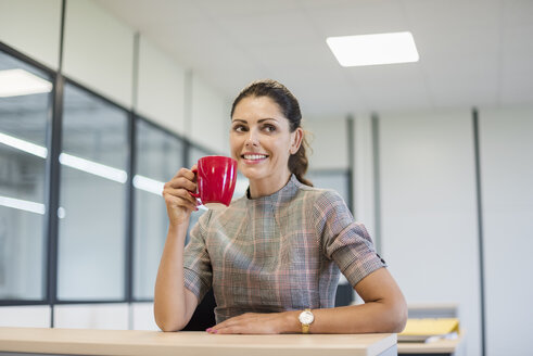Hübsche Büroangestellte sitzt am Schreibtisch mit roter Kaffeetasse - DIGF05275