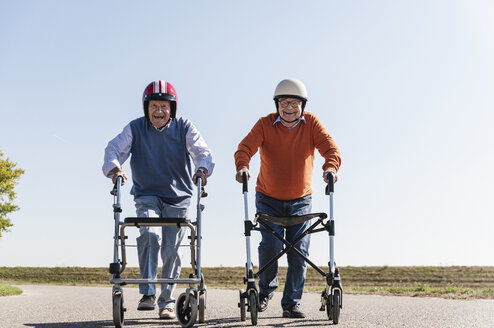 Two old friends wearing safety helmets, competing in a wheeled walker race - UUF15541