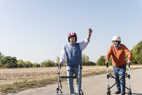 Two old friends wearing safety helmets, competing in a wheeled walker race - UUF15542