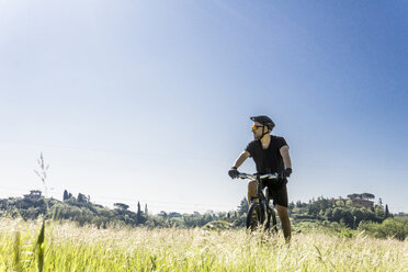 Junger Mann mit Mountainbike auf grasbewachsenem Feld gegen den klaren Himmel an einem sonnigen Tag - CAVF49986