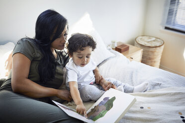 Niedlicher Sohn, der auf ein Bilderbuch zeigt, während er neben seiner Mutter auf dem Bett sitzt - CAVF50017