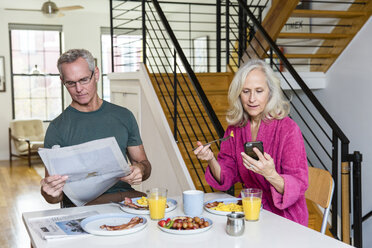 Mann liest Zeitung, während Frau am Esstisch ein Smartphone benutzt - CAVF50062