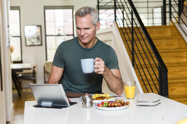 Älterer Mann hält Kaffeetasse, während er einen Tablet-Computer auf dem Esstisch zu Hause benutzt - CAVF50063