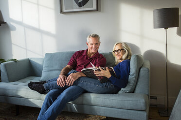 Smiling couple reading book while resting on sofa in living room at home - CAVF50064