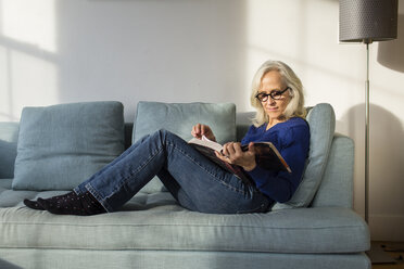 Full length of senior woman reading book while resting on sofa at home - CAVF50065