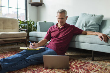 Ernster reifer Mann, der einen Laptop benutzt, während er auf dem Teppich neben dem Sofa im Wohnzimmer sitzt - CAVF50067