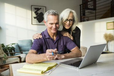 Lächelnde Frau, die auf einen Laptop schaut, während ein Mann eine Kreditkarte im Wohnzimmer hält - CAVF50073