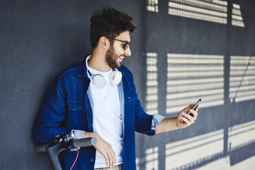 Lächelnder junger Mann mit Elektroroller schaut auf sein Handy im Freien - BSZF00763