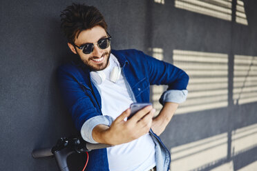 Porträt eines lächelnden jungen Mannes mit Elektroroller, der auf sein Mobiltelefon schaut - BSZF00764