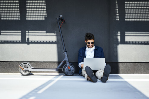 Relaxed young man sitting on the ground besides his electric scooter using laptop - BSZF00769