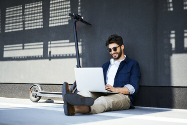 Smiling young man sitting on the ground besides his electric scooter using laptop - BSZF00770