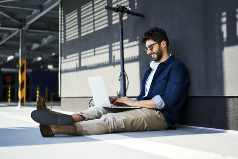 Smiling young man with electric scooter sitting on the ground using laptop stock photo