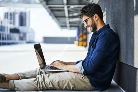 Smiling young man sitting on the ground using laptop stock photo