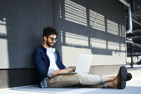 Lächelnder junger Mann, der auf dem Boden sitzt und einen Laptop benutzt, lizenzfreies Stockfoto