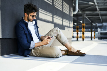 Lächelnder junger Mann, der auf dem Boden sitzt und ein Smartphone benutzt - BSZF00778