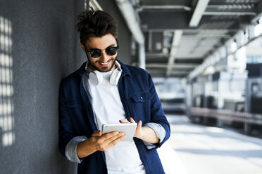 Smiling young man leaning against wall using digital tablet - BSZF00781