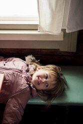 Porträt eines lächelnden kleinen Mädchens, das auf einer Holzbank liegt und einen Anorak im Haus trägt - PSIF00116