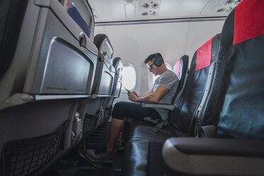 Mann im Flugzeug, mit Smartphone und Kopfhörern - KKAF02453
