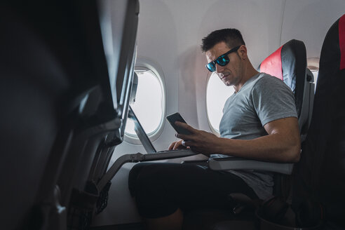 Mann im Flugzeug, mit Smartphone und Laptop - KKAF02459