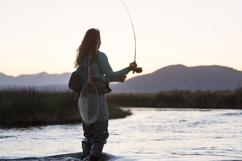 Seitenansicht einer jungen Frau beim Fliegenfischen im Owens River mit Blick auf die Berge bei Sonnenuntergang - CAVF50119