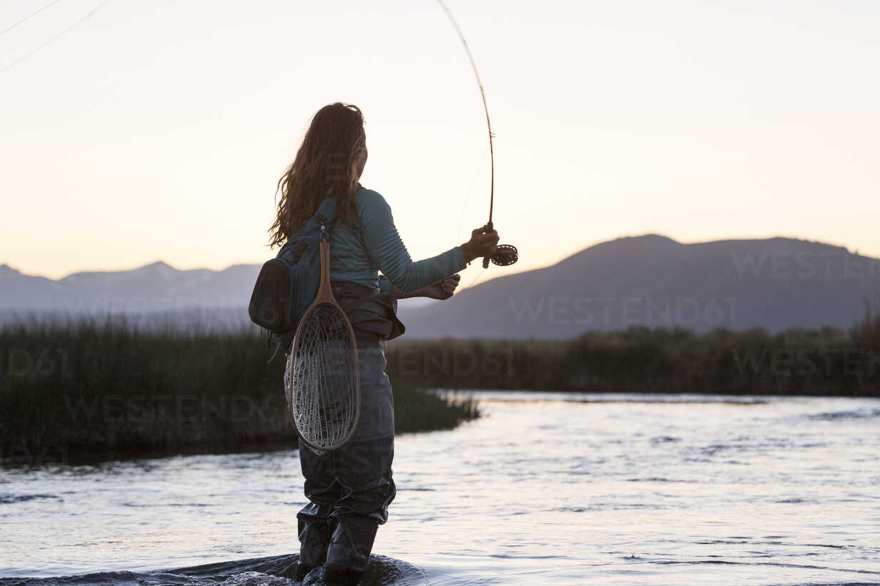 Side view of young woman fly-fishing while standing in Owens River against  mountains during sunset stock photo
