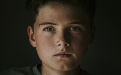 Close-up portrait of serious boy standing in darkroom at home - CAVF50126