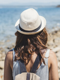 Rückansicht einer Frau mit Rucksack, die an einem sonnigen Tag am felsigen Strand steht und die Aussicht betrachtet - CAVF50184