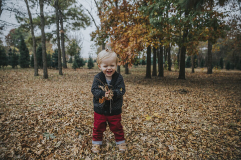 Porträt eines glücklichen kleinen Jungen, der mit Herbstblättern spielt, während er im Park steht, lizenzfreies Stockfoto