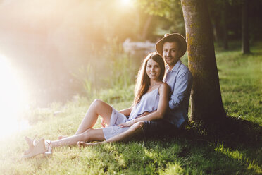 Portrait of couple sitting on grassy field by tree at park - CAVF50247