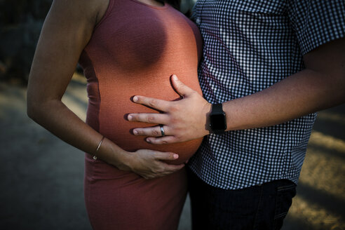 Mittelteil des Ehemanns, der den Bauch seiner schwangeren Frau berührt, während er bei Sonnenuntergang im Park steht - CAVF50260