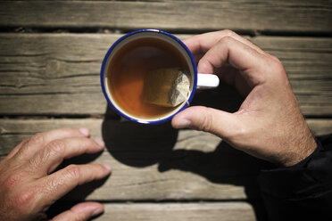 Abgeschnittene Hände eines Mannes, der Tee in einer Tasse auf einem Holztisch hält - CAVF50272