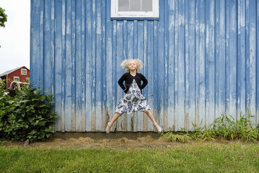 Porträt eines niedlichen Mädchens mit Händen auf der Hüfte, das auf einer Wiese gegen eine Mauer springt - CAVF50298