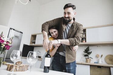 Portrait of happy couple with bottle of red wine in the kitchen - KMKF00574
