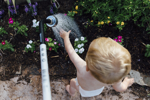 Rückansicht des hemdlosen Babyjungen, der mit Wasser spielt, während er bei den Pflanzen im Garten steht, lizenzfreies Stockfoto