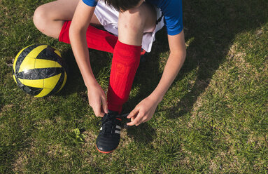 Hoher Blickwinkel des Jungen Schnürsenkel binden, während durch Fußball auf Rasenplatz sitzen - CAVF50326