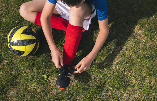 High angle view of boy tying shoelace while sitting by soccer ball on grassy field - CAVF50326
