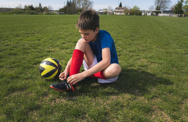 Full length of boy tying shoelace while sitting by soccer ball on grassy field - CAVF50327