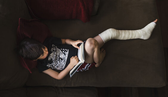 High angle view of boy with broken leg reading book while lying on sofa at home - CAVF50334