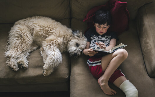 High angle view of boy with fractured leg reading book while lying by dog on sofa at home - CAVF50335