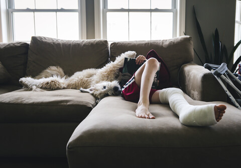 Niedriger Ausschnitt eines Jungen mit gebrochenem Bein, der ein Buch liest, während er mit seinem Hund zu Hause auf dem Sofa liegt, lizenzfreies Stockfoto