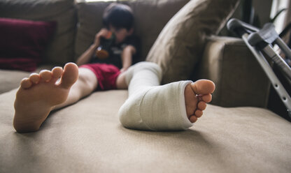 Junge mit gebrochenem Bein sitzt zu Hause auf dem Sofa - CAVF50337