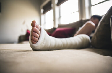 Close-up of boy with fractured leg sitting on sofa at home - CAVF50338